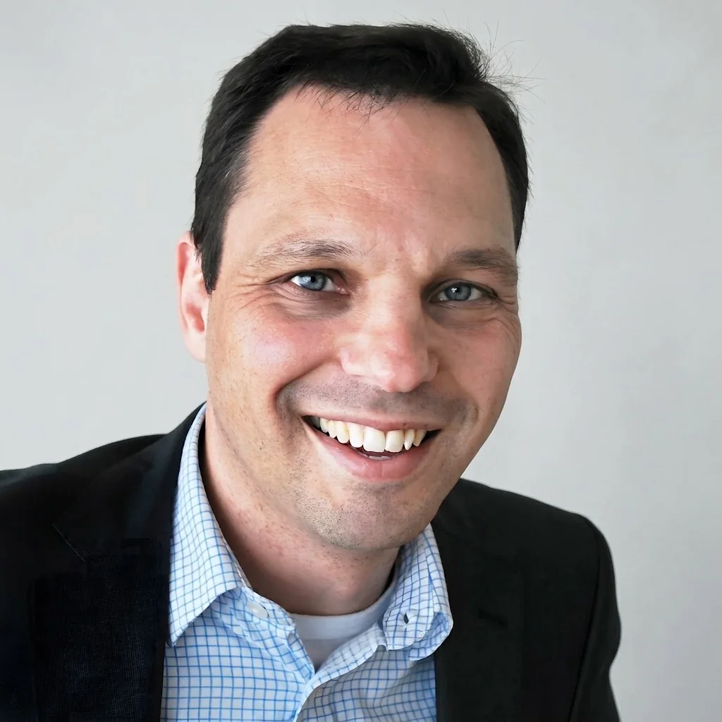 A smiling man in a suit and checkered shirt against a plain background.