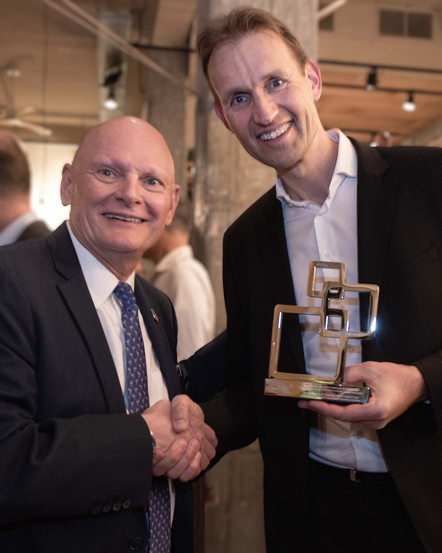 Two men in suits smiling and shaking hands, one holding a trophy.