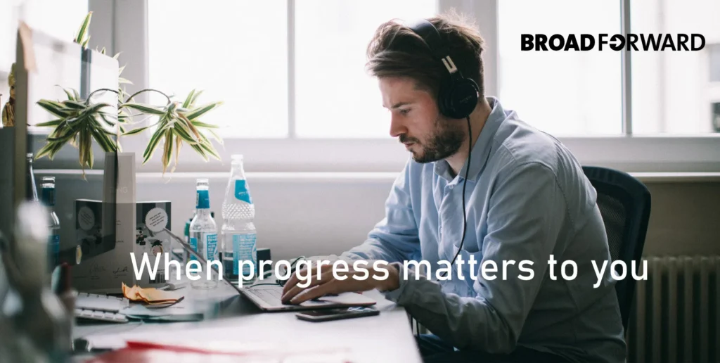 A focused developer wearing headphones works on a laptop at a desk with a plant and water bottles, accompanied by the text "When progress matters to you" and the logo "BroadForward."