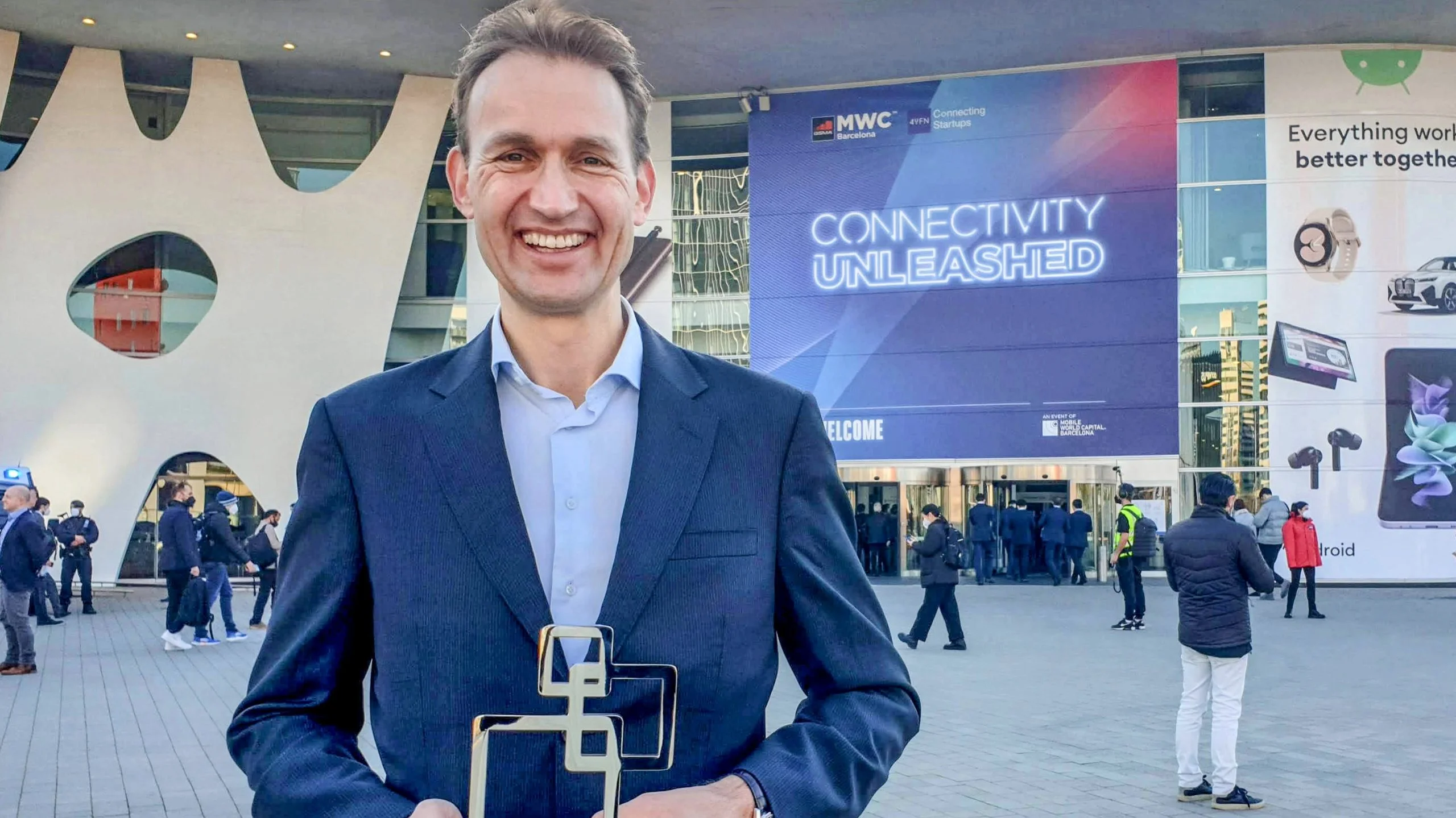 A man in a suit holding an award in front of a technology conference building with a "Connectivity Unleashed" banner.