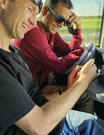 Two men sitting in a vehicle, smiling and looking at a phone.