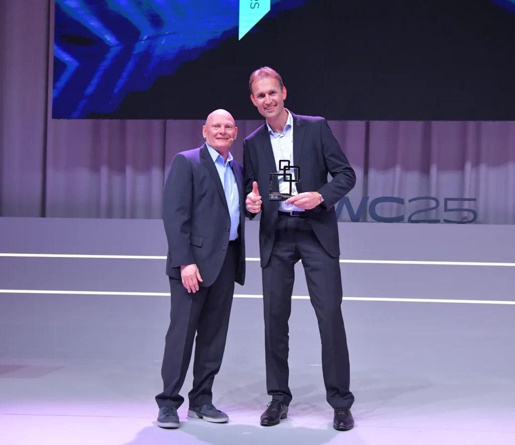Two men in suits on stage at the MWC25 award ceremony, with one holding a trophy and giving a thumbs up.