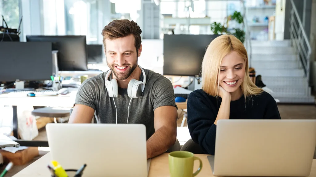 Two colleagues smiling and working on laptops in a modern office.
