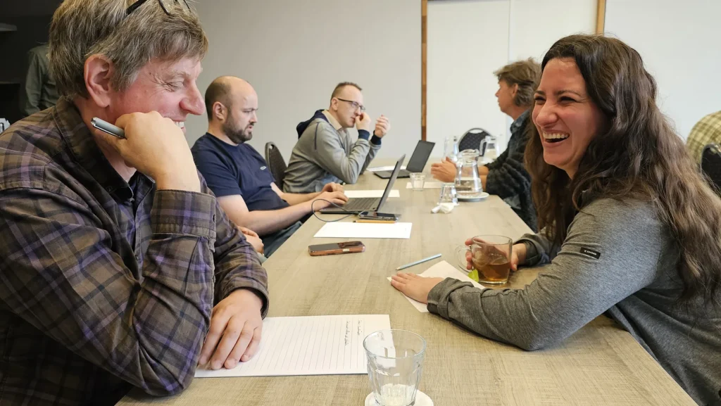 Colleagues laughing together during a workshop session at a table with notebooks and laptops.